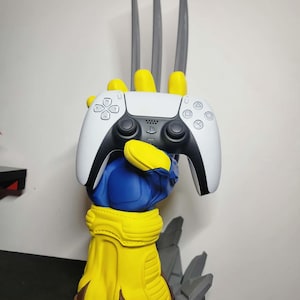Puede incluir: Un soporte para mando con forma de guante amarillo y azul que presenta tres garras largas, grises y puntiagudas que se extienden hacia arriba. El soporte sostiene un mando de juego inalámbrico blanco y negro con botones, joysticks y panel táctil.