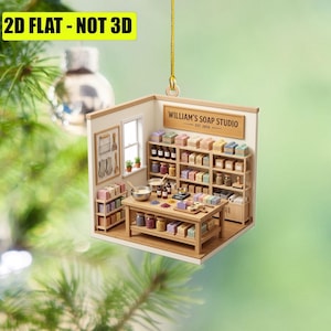 May include: A flat, two-dimensional hanging ornament depicting a miniature soap studio interior. The scene features wooden shelves filled with colorful soap bars, a central workbench with mixing bowls, and a sign reading WILLIAM'S SOAP STUDIO EST. 2026.