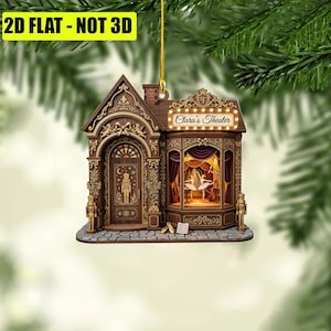 May include: A flat, two-dimensional hanging ornament shaped like a theater building with brown wood-toned details. The facade features a door with a nutcracker graphic, a stage with two ballerinas, and a sign reading Clara's Theater.