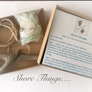 Può includere: Una scatola di cartone marrone su uno sfondo bianco. La scatola contiene un kit per realizzare un acchiappasole. Il kit include un pezzo di legno galleggiante, spago e vetro di mare. La scatola contiene istruzioni su come realizzare l'acchiappasole. Il testo sulla scatola recita "Shore Things... Make Your Own Sun Catcher Instructions".