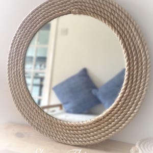 May include: A round mirror with a thick rope frame made of natural jute. The mirror is hanging on a wall and reflects a room with cushions on a sofa.