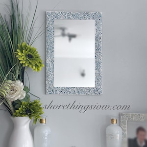 May include: A rectangular mirror with a mosaic tile frame in shades of blue and white hangs on a light grey wall. Below, white vases and decorative bottles with gold accents sit on a shelf, alongside a small mosaic piece and a framed photo.