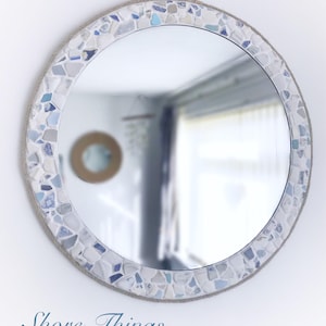May include: A round mirror with a white and blue mosaic frame made of sea glass and rope. The mirror is hanging on a white wall. "Shore Things..." is written in cursive below the mirror.