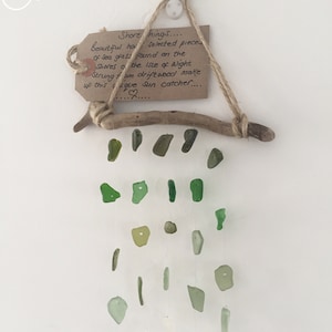 Puede incluir: Un atrapasol de madera flotante con piezas de vidrio de mar verde colgando de él. El atrapasol está colgado de un trozo de cuerda y una pequeña etiqueta marrón con el texto "Shore things... Beautiful hand selected pieces of sea glass found on the shores of the Isle of Wight. String from driftwood make up this unique sun catcher..."