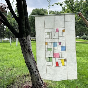 Pode incluir: Uma tapeçaria retangular de tecido branco sujo suspensa por uma haste horizontal. O centro apresenta uma grade de linhas costuradas formando várias formas geométricas, algumas preenchidas com retalhos translúcidos nas cores rosa, azul, amarelo, verde e vermelho.