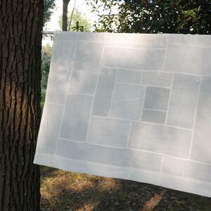 K&ouml;nnte beinhalten: Ein rechteckiges wei&szlig;es Stoffpaneel mit einem geometrischen Patchwork-Design aus verschieden gro&szlig;en Rechtecken und Quadraten. Das halbtransparente Textil zeigt ein gitterartiges Muster aus gen&auml;hten N&auml;hten, das eine strukturierte Komposition bildet.