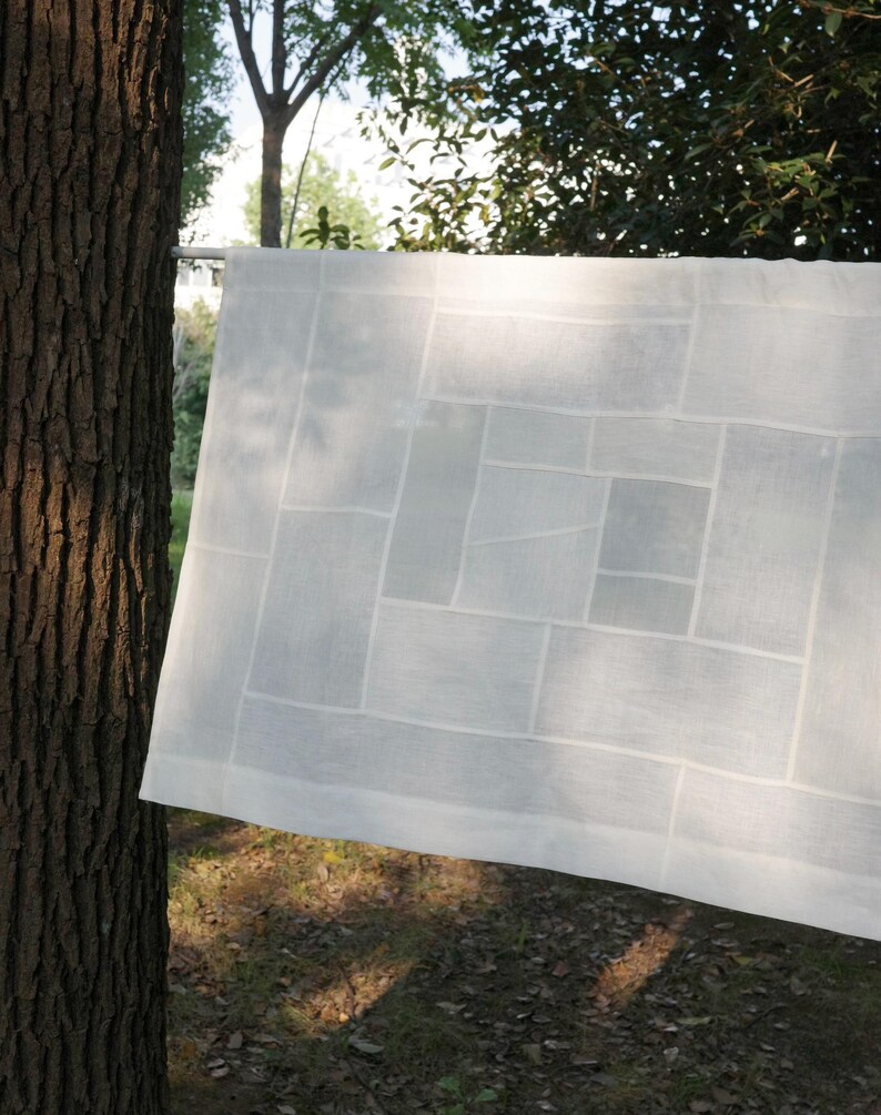K&ouml;nnte beinhalten: Ein rechteckiges wei&szlig;es Stoffpaneel mit einem geometrischen Patchwork-Design aus verschieden gro&szlig;en Rechtecken und Quadraten. Das halbtransparente Textil zeigt ein gitterartiges Muster aus gen&auml;hten N&auml;hten, das eine strukturierte Komposition bildet.