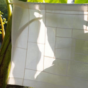 K&ouml;nnte beinhalten: Ein rechteckiges, cremewei&szlig;es Stoffpaneel mit einem geometrischen Gittermuster aus gen&auml;hten N&auml;hten. Das halbtransparente Textil zeigt eine Reihe verschiedener rechteckiger und quadratischer Abschnitte, die zu einer strukturierten, linearen Komposition zusammengef&uuml;gt sind.