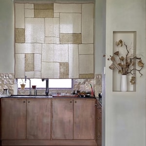 May include: A rectangular window shade featuring a patchwork design of cream and tan fabric panels with visible dark stitching. The shade hangs above a kitchen sink with wooden cabinets below and a white vase with dried branches in a wall niche.