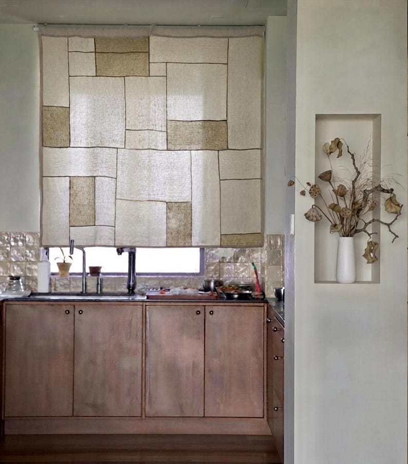 Puede incluir: Una cortina de ventana rectangular con un dise&ntilde;o de patchwork de paneles de tela color crema y tostado con costuras oscuras visibles. La cortina cuelga sobre un fregadero de cocina con armarios de madera y un jarr&oacute;n blanco con ramas.