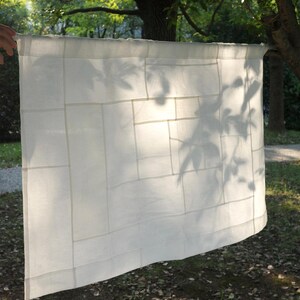 K&ouml;nnte beinhalten: Ein rechteckiges Paneel aus wei&szlig;em Stoff mit einem Patchwork-Design aus verschieden gro&szlig;en, zusammengen&auml;hten Rechtecken. Das halbtransparente Material weist ein sichtbares Raster aus N&auml;hten und einen Tunnelzug zur Aufh&auml;ngung auf.