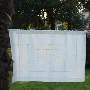 K&ouml;nnte beinhalten: Ein rechteckiges wei&szlig;es Stoffpaneel mit einem geometrischen Patchwork-Design, das aus verschieden gro&szlig;en rechteckigen und quadratischen Segmenten besteht, die mit feinen, sichtbaren N&auml;hten zu einem Gittermuster verbunden sind.