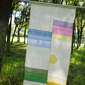 May include: A rectangular, semi-sheer white fabric panel featuring a geometric patchwork design. It includes horizontal bands of blue, green, yellow, pink, and purple, along with a solid yellow circle positioned on the right side of the panel.