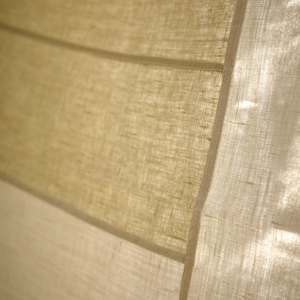 Puede incluir: Una cortina de tela semitransparente con una textura tejida visible en tonos beige claro y bronceado. El material presenta paneles horizontales unidos por costuras finas y rectas, permitiendo que la luz pase a trav&eacute;s del tejido fibroso y transl&uacute;cido.