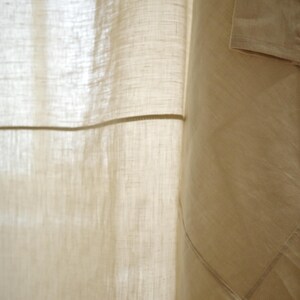 Puede incluir: Una cortina de color beige hecha de una tela semitransparente y texturizada con un tejido visible. La cortina presenta un patr&oacute;n de cuadr&iacute;cula creado por costuras horizontales y verticales, con un panel beige s&oacute;lido m&aacute;s oscuro a la derecha.