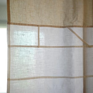 Könnte beinhalten: Ein halbtransparenter Vorhang mit einer Farbpalette in Hellbeige und Off-White. Der Stoff weist eine sichtbare Webstruktur mit horizontalen und vertikalen Bändern in einem dunkleren Beigeton auf, die ein gitterartiges Muster auf der Oberfläche bilden.