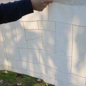 K&ouml;nnte beinhalten: Ein rechteckiges, cremewei&szlig;es Stoffpaneel mit einem geometrischen Gittermuster aus d&uuml;nnen, halbtransparenten vertikalen und horizontalen Linien. Das Textil hat eine glatte, leichte Optik und wird gegen das Licht gehalten.