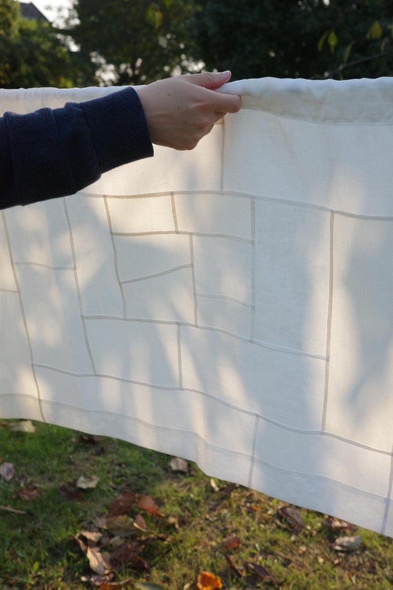 K&ouml;nnte beinhalten: Ein rechteckiges, cremewei&szlig;es Stoffpaneel mit einem geometrischen Gittermuster aus d&uuml;nnen, halbtransparenten vertikalen und horizontalen Linien. Das Textil hat eine glatte, leichte Optik und wird gegen das Licht gehalten.