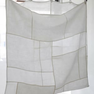 Puede incluir: Una pieza textil cuadrada de color blanco con un patrón de cuadrícula formado por finas costuras de color beige claro que dividen la tela en varias secciones rectangulares y cuadradas. La tela cuelga de una barra metálica con cinco clips.