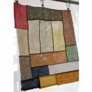 May include: A rectangular textile wall hanging composed of various linen-like fabric patches in shades of red, olive green, brown, yellow, tan, blue, and black. The pieces are joined by thin brown seams, creating a grid-like patchwork design.