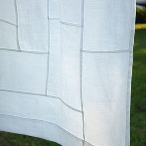 K&ouml;nnte beinhalten: Ein wei&szlig;er Vorhang aus halbtransparentem Stoff mit einem gitterartigen Muster aus vertikalen und horizontalen N&auml;hten. Das helle Textil weist eine sichtbare Webstruktur auf und wird vom Sonnenlicht beleuchtet, was weiche Schatten an den N&auml;hten erzeugt.
