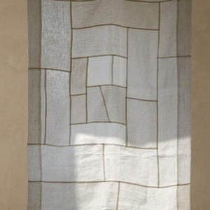 May include: A rectangular linen curtain featuring a grid of light beige and off-white panels. The fabric is semi-sheer, allowing light to pass through, and is suspended from a thin wire by three wooden clothespins at the top edge.