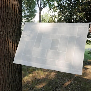 May include: A rectangular white fabric panel featuring a geometric patchwork design of various sized rectangles. The semi-sheer textile is suspended horizontally from a thin white rod, displaying a grid-like pattern of stitched seams.