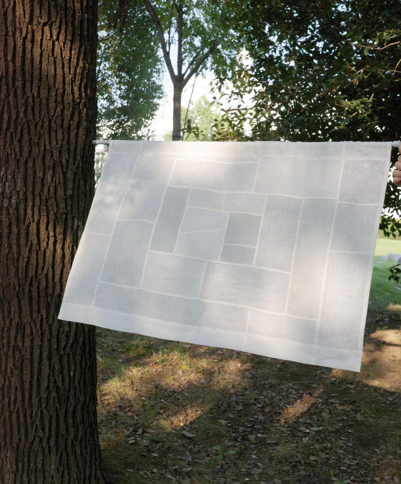 K&ouml;nnte beinhalten: Ein rechteckiges wei&szlig;es Stoffpaneel mit einem geometrischen Patchwork-Design aus verschieden gro&szlig;en Rechtecken. Das halbtransparente Textil ist horizontal an einer d&uuml;nnen wei&szlig;en Stange befestigt und zeigt N&auml;hte.