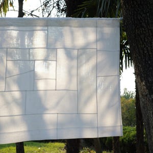 K&ouml;nnte beinhalten: Ein wei&szlig;es, rechteckiges Stoffpaneel mit einem Patchwork-Design aus verschieden gro&szlig;en Rechtecken und Quadraten. Das halbtransparente Material ist mit sichtbaren N&auml;hten zusammengesetzt, die die einzelnen Teile verbinden und ein Gittermuster bilden.