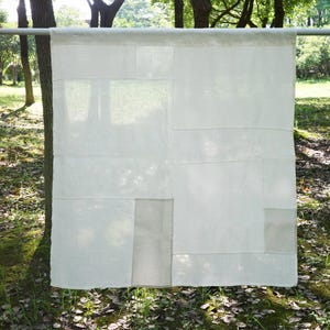 Puede incluir: Un panel de tela rectangular de color blanco roto con un diseño de patchwork compuesto por cuadrados y rectángulos de varios tamaños. El tejido está formado por múltiples segmentos de material claro similar al lino con costuras visibles.