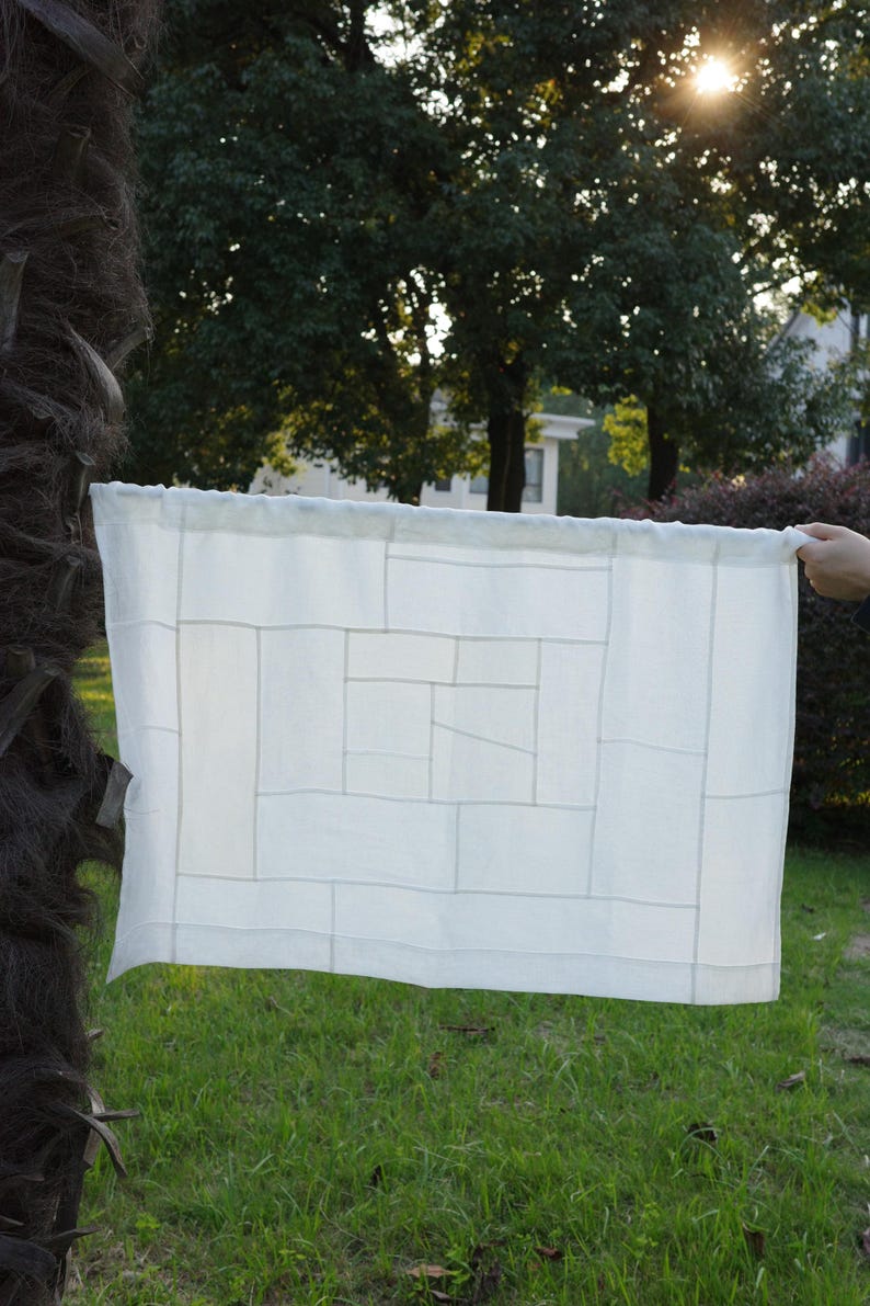 K&ouml;nnte beinhalten: Ein rechteckiger, cremewei&szlig;er Stoffvorhang mit einem geometrischen Patchwork-Design, das aus verschiedenen zusammengen&auml;hten rechteckigen Paneelen besteht. Der Stoff hat einen Stangendurchzug an der Oberkante und wirkt halbtransparent mit sichtbaren N&auml;hten.
