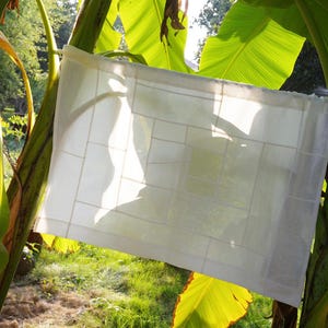 K&ouml;nnte beinhalten: Ein rechteckiges Paneel aus wei&szlig;em Stoff mit einem Gitter aus gen&auml;hten Linien, die ein geometrisches Muster aus verschiedenen rechteckigen Formen bilden. Das halbtransparente Textil ist aufgeh&auml;ngt, sodass Licht durch den Stoff f&auml;llt.