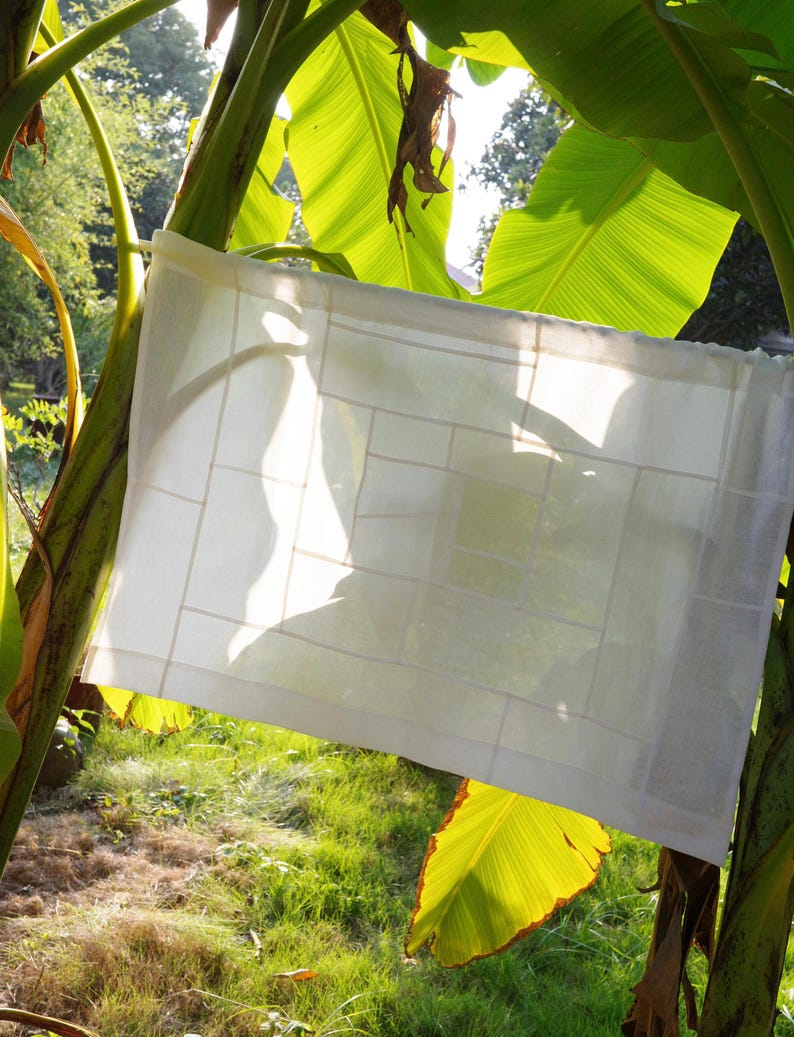K&ouml;nnte beinhalten: Ein rechteckiges Paneel aus wei&szlig;em Stoff mit einem Gitter aus gen&auml;hten Linien, die ein geometrisches Muster aus verschiedenen rechteckigen Formen bilden. Das halbtransparente Textil ist aufgeh&auml;ngt, sodass Licht durch den Stoff f&auml;llt.