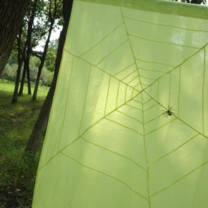 May include: A square, light lime green fabric panel featuring a dark green, printed spider web pattern that radiates from the centre. A small, dark, multi-legged spider figure is attached to the web, positioned slightly off-centre on the fabric.