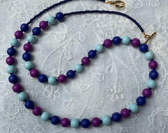May include: A beaded necklace featuring a repeating pattern of round dark blue, light blue, and purple beads. The necklace includes a thin strand of tiny dark blue seed beads and is secured with a gold-toned metal toggle clasp closure.