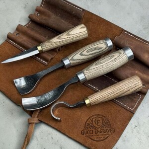 May include: A set of four woodworking tools with light-colored wood handles and metal blades, including a straight knife, two gouges, and a hook knife, arranged on a brown leather tool roll embossed with the text Gucci Engraved Handcrafted Woodwork.