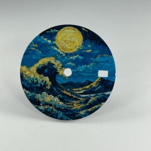 Puede incluir: Una esfera de reloj circular con una escena pintada de una gran ola rompiendo en tonos azul profundo y dorado bajo una luna amarilla brillante. La esfera tiene un orificio central circular y una pequeña abertura rectangular a la derecha.
