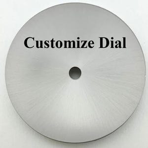 Puede incluir: Un dial circular de metal plateado con una textura radial cepillada que se extiende desde el centro hasta el borde exterior. Presenta un pequeño orificio circular centrado. El texto "Customize Dial" está impreso en negro en la mitad superior.
