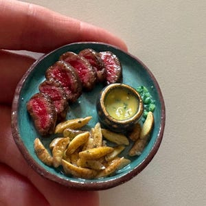May include: A miniature ceramic plate featuring a serving of sliced red meat, a pile of golden-brown potato wedges, and a small dark bowl filled with yellow sauce. The plate has a teal center with a dark brown rim, presenting a detailed food scene.