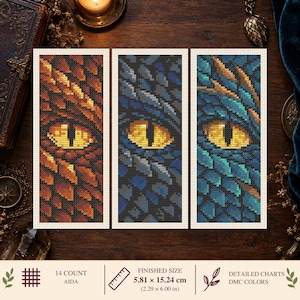 May include: Three rectangular cross-stitch patterns featuring dragon eyes surrounded by scales in orange, dark blue, and teal. The patterns are printed on white fabric. A ruler icon indicates a finished size of 2.29 by 6.00 inches on 14 count Aida.