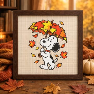 May include: A framed cross-stitch embroidery featuring a white cartoon dog with black ears holding a colourful umbrella made of orange, red, and yellow autumn leaves. Several individual fall leaves are scattered around the dog on a light fabric.