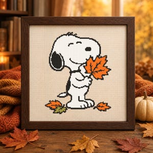 May include: A framed cross-stitch embroidery featuring a white beagle with black ears holding a large orange maple leaf. The design is stitched on a light beige Aida cloth, depicting the character standing among scattered orange and green leaves.