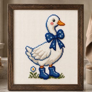 May include: A framed cross-stitch embroidery featuring a white goose with a blue polka-dot bow around its neck. The goose wears blue rain boots and stands on a small patch of green grass with a single white flower, all set in a dark wood frame.