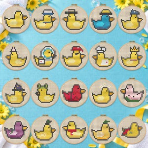 Puede incluir: Una colección de veinte bastidores de bordado circulares de madera, cada uno con un diseño único de punto de cruz pixel art de un patito de goma. Los patos presentan varios accesorios como sombreros, coronas y disfraces en diversos colores.