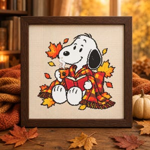 May include: A framed cross-stitch embroidery featuring a white cartoon dog with a black ear, wrapped in a red and orange plaid blanket. The dog holds a steaming orange mug and an open red book, surrounded by scattered yellow and orange maple leaves.