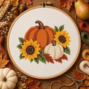 Può includere: Un telaio da ricamo circolare in legno contenente un motivo a punto croce raffigurante una grande zucca arancione, una zucca bianca più piccola e due girasoli gialli con foglie verdi su uno sfondo di tessuto bianco con trama a griglia.