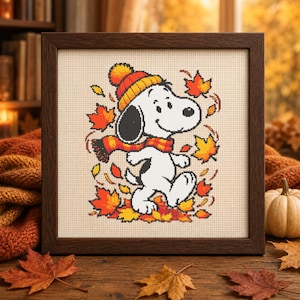 May include: A framed cross-stitch embroidery featuring a white dog with black ears wearing an orange and yellow striped knit hat and a matching plaid scarf, walking amidst falling red, orange, and yellow autumn maple leaves on a beige fabric.