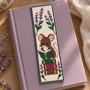May include: A rectangular cross-stitch bookmark on a cream background featuring a brown mouse with pink ears reading a book while sitting on a stack of three books. The design includes purple lavender sprigs and green leaves at the top and bottom.