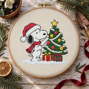 Pode incluir: Um bastidor de bordado circular de madeira contendo um padrão de ponto cruz de um beagle branco com gorro e cachecol de Pai Natal, decorando uma árvore de Natal verde com enfeites vermelhos, amarelos e dourados e dois presentes.