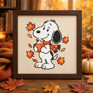 May include: A framed cross-stitch embroidery featuring a white cartoon dog with a black ear, wearing an orange and red plaid scarf. The dog holds an orange mug with whipped cream, surrounded by falling orange and red maple leaves on fabric.